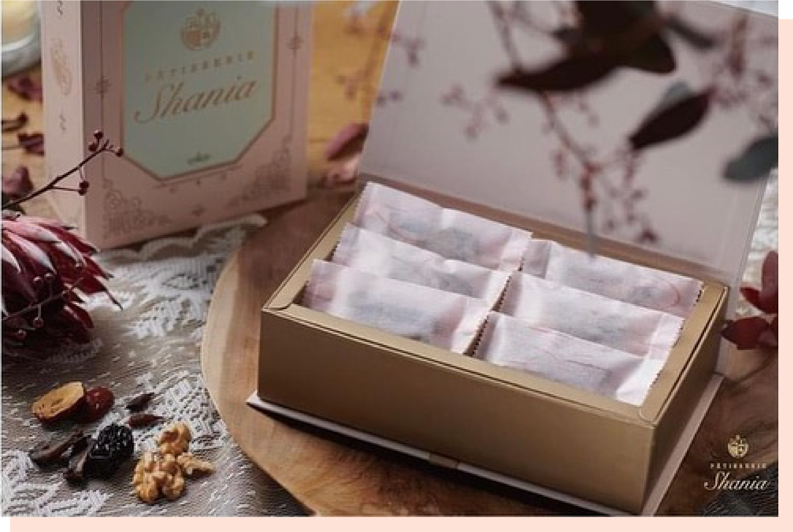 PÂTISSERIE品牌的粉色禮盒打開，內裝有用粉色紙包裹的商品，盒蓋上寫有“Thank You”，背景是木質桌面，旁邊散落著些許秋季裝飾。