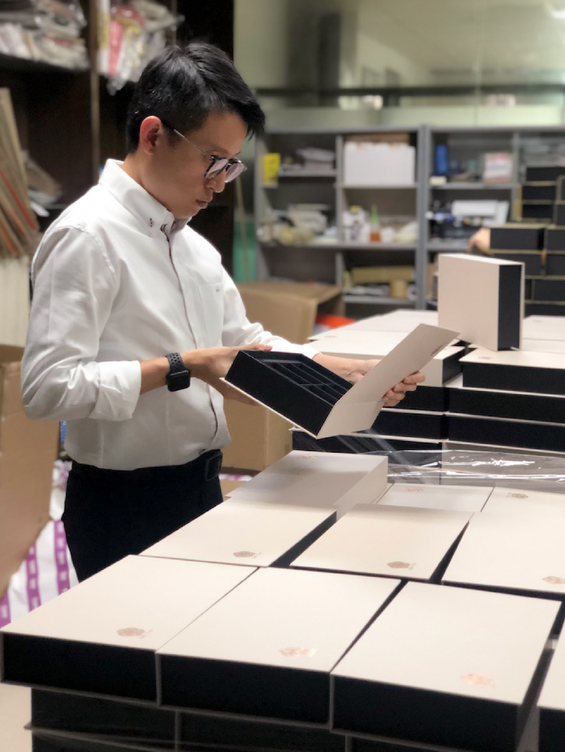 一張在包裝工作區的照片，一位穿著白色襯衫和眼鏡的男士正在認真檢查一個打開的白色禮品盒。背景中可見排列整齊的多個相同的禮品盒，表面帶有品牌標誌
