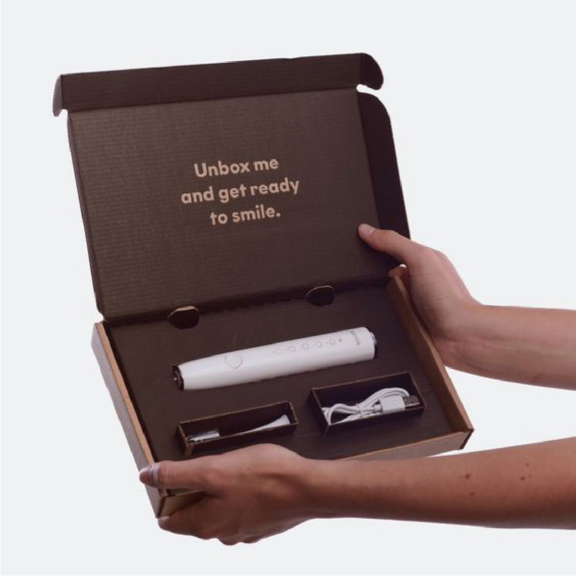 在灰色的背景下，一位人士正打開深啡色的郵寄盒，盒內的蓋寫有"Unbox me and get ready to smile"的字眼。