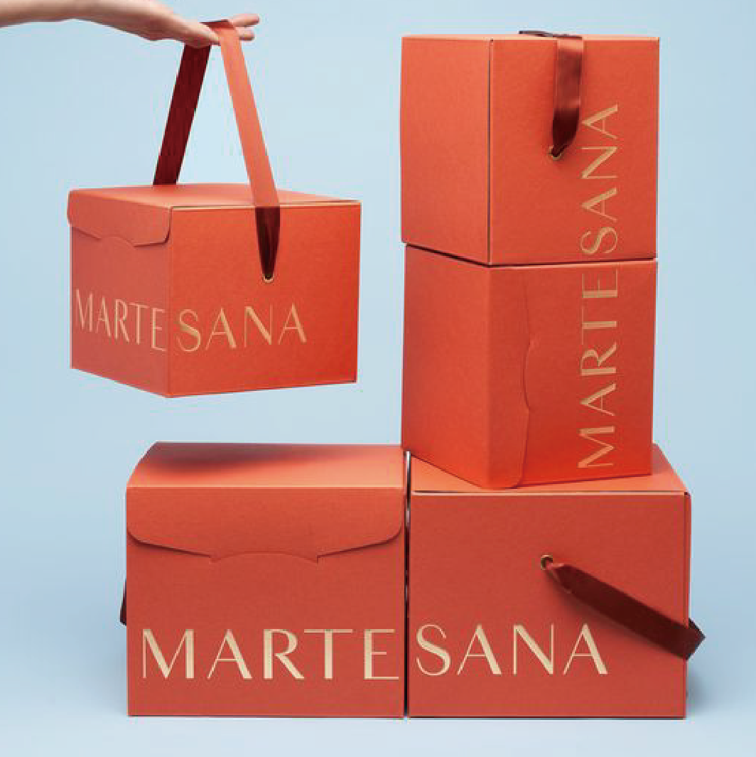 有五個橙色的盒，左邊下方擺放了一個，上方有一隻手提起著盒子，右邊有三個橙色盒疊起。最底下兩個盒子合拉後帶有"MARTESANA"的字樣。