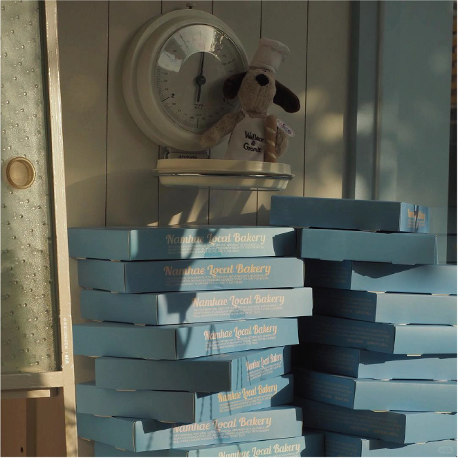 A stack of neatly stacked ocean blue boxes with the words 'Namhae Local Bakery' printed on them, topped with a doll dog wearing a baker's hat, creates a warm and cosy atmosphere.
