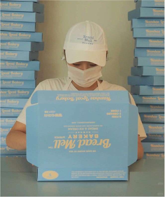 Uniformed bakery staff wearing branded hats look intently at the ocean blue boxes with the words 'Bread Mell Bakery' printed on them. In the background is a stack of boxes with 'Namhae Local Bakery' printed on them.