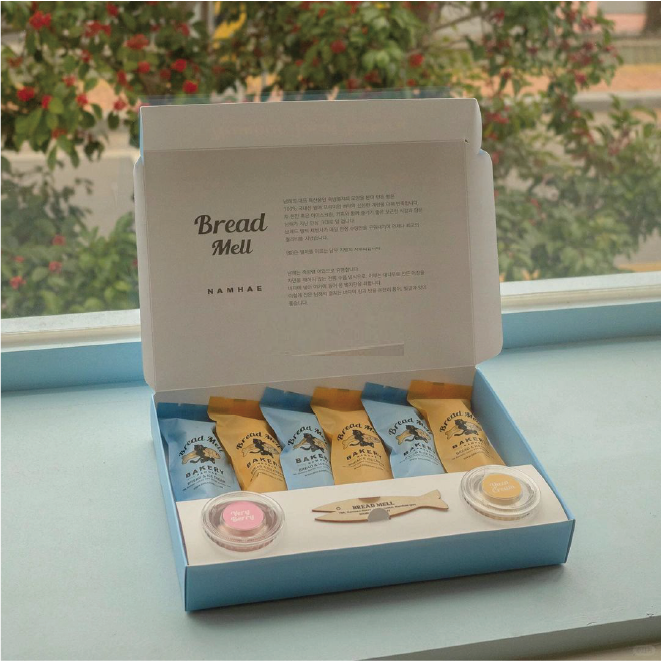 An ocean blue 'Bread Mell Bakery' bakery box with the brand name and description printed on the lid. Inside the box are several beautifully wrapped buns and two small bottles of jam.