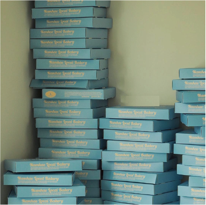 A mountain of ocean blue bakery boxes, each with 'Namhae Local Bakery' clearly printed in white lettering. The boxes are neatly stacked in front of a light-coloured wall.