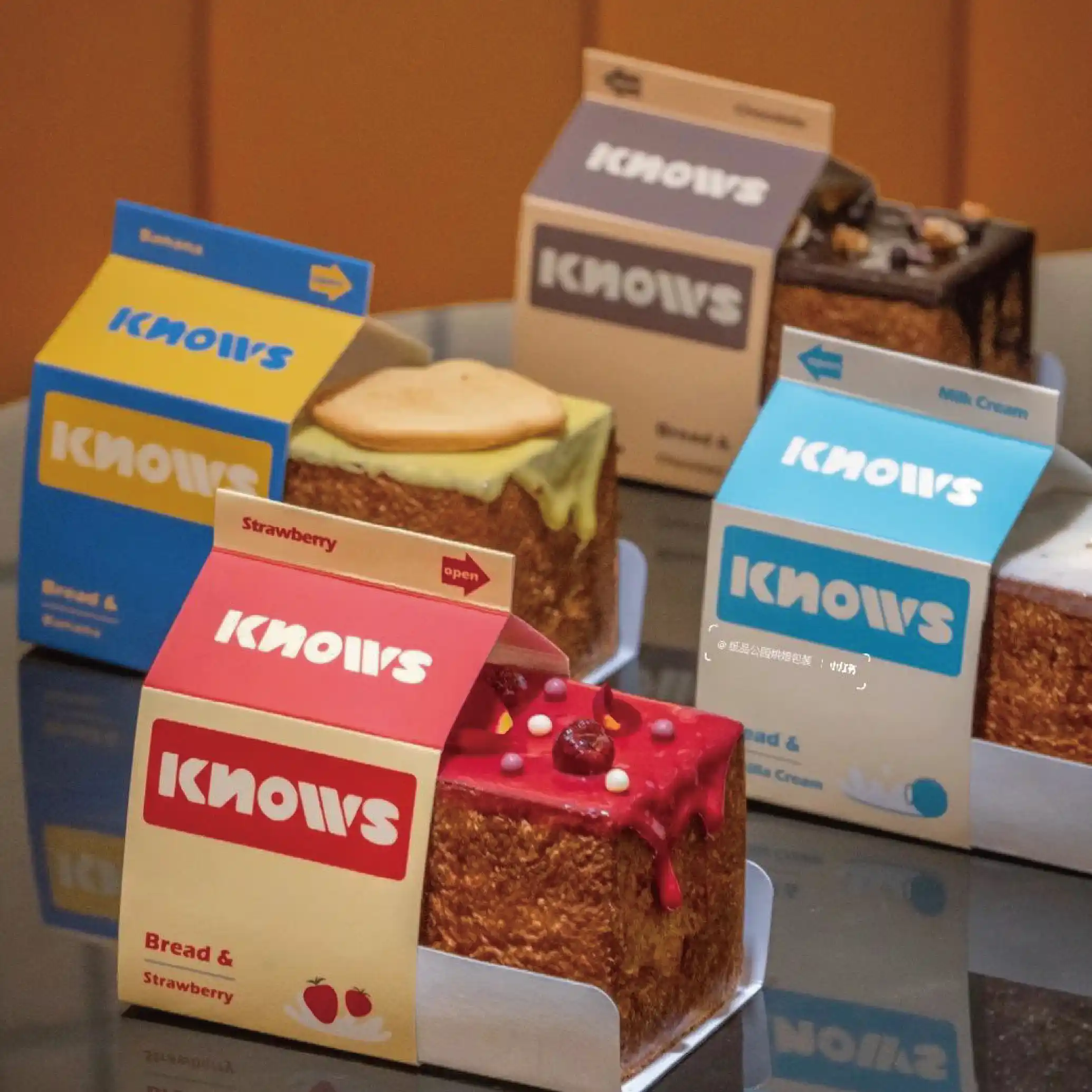 A series of brightly coloured cake boxes with a red and yellow box in the foreground and an orange background with the brand 'KNOWS' logo and labels for the different cake flavours.