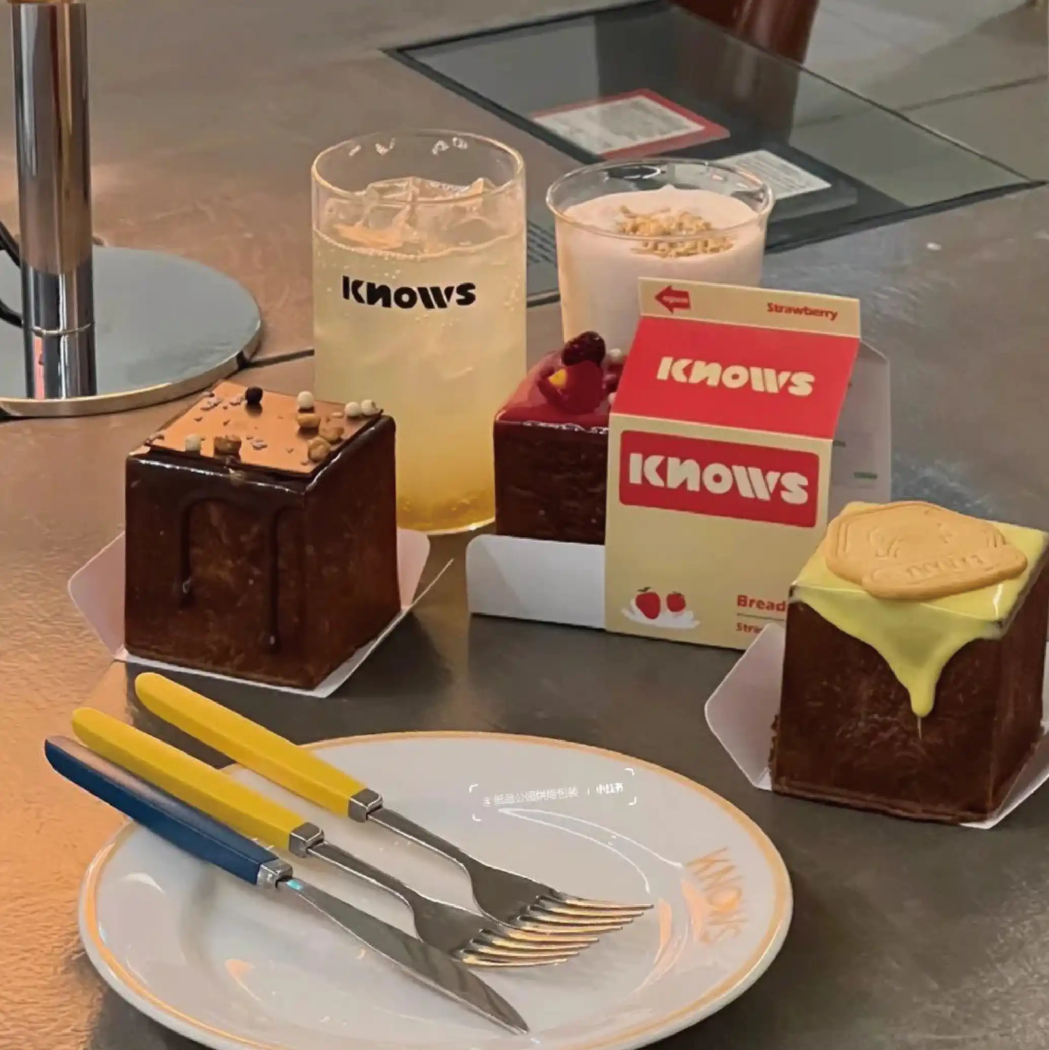 On a dining table, there is a clear drinking glass and two cakes, one strawberry flavoured and the other banana flavoured, next to a cake knife and fork.