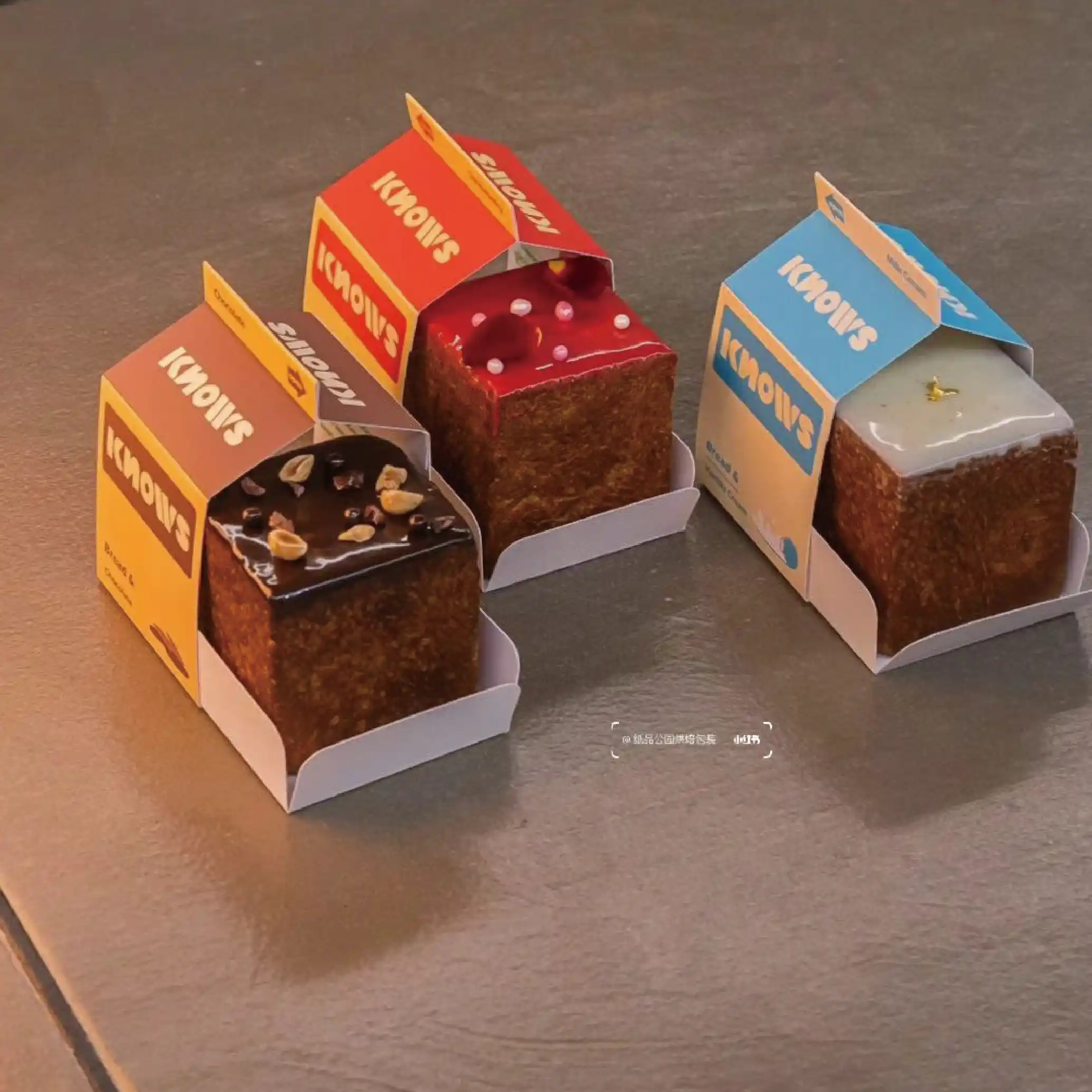 A range of different flavoured cakes and their corresponding 'KNOWS' branded boxes, each with unique decorations such as chocolate chips and jam.
