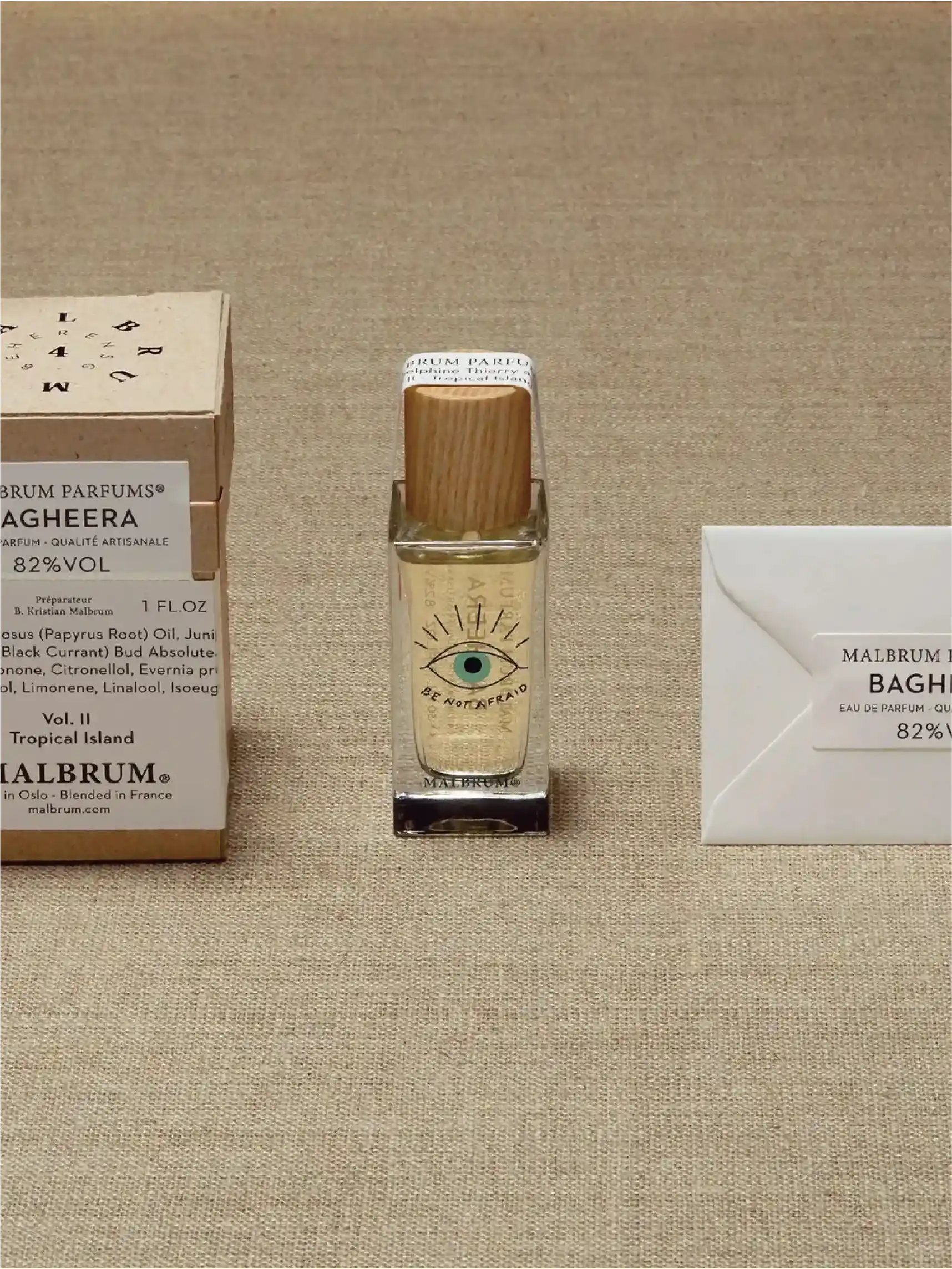 A clear bottle of perfume with a wooden lid sits next to an open brown cardboard box with a white card with the name of the perfume 'WILDFIRE' printed on it in the background.