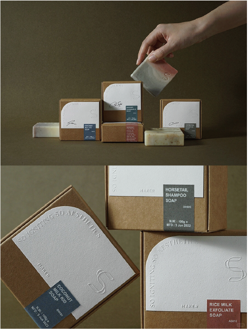 Multiple soap boxes are lined up on a brown background with a hand picking up one of the bars of soap. Each box has the words "SO GIFTING SO AESTHETICS" and "MAKER" and a label describing the type of soap.