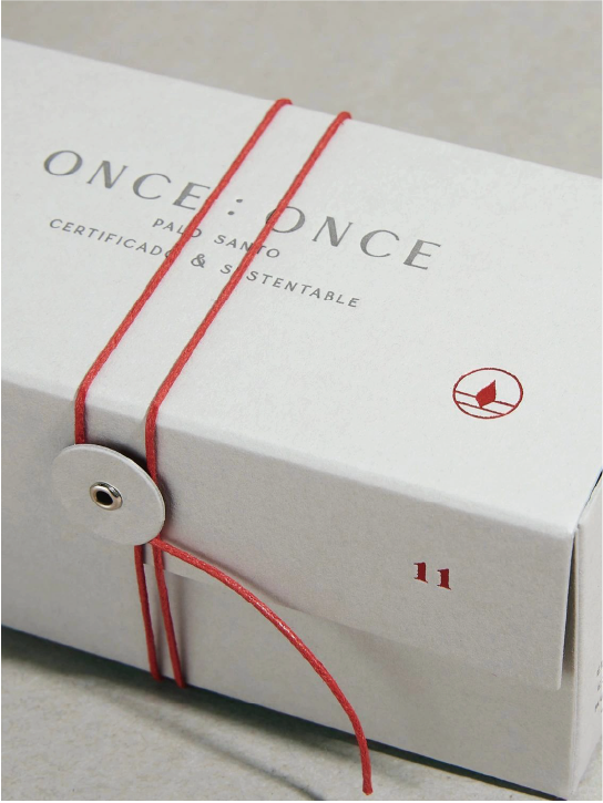 The white box is printed with "ONCE : ONCE PALO SANTO CERTIFICADO & SUSTENTABLE", with the brand's logo next to it, decorated with a red cord and tied with a round label.