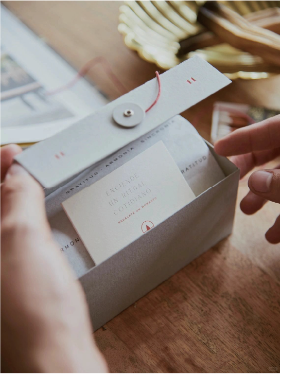 The hand is opening the grey box containing the card, the lid of which is tied with a red cord with a snap closure, and the card has the brand logo and text on it.