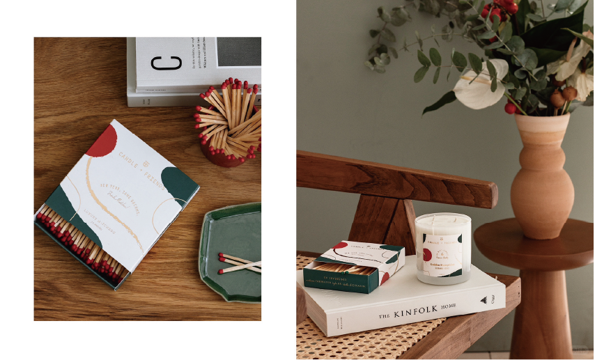 Matchboxes and candles are placed on a wooden table with the words "CANDLE & FRIENDS" on the box, next to a book and a vase of flowers.