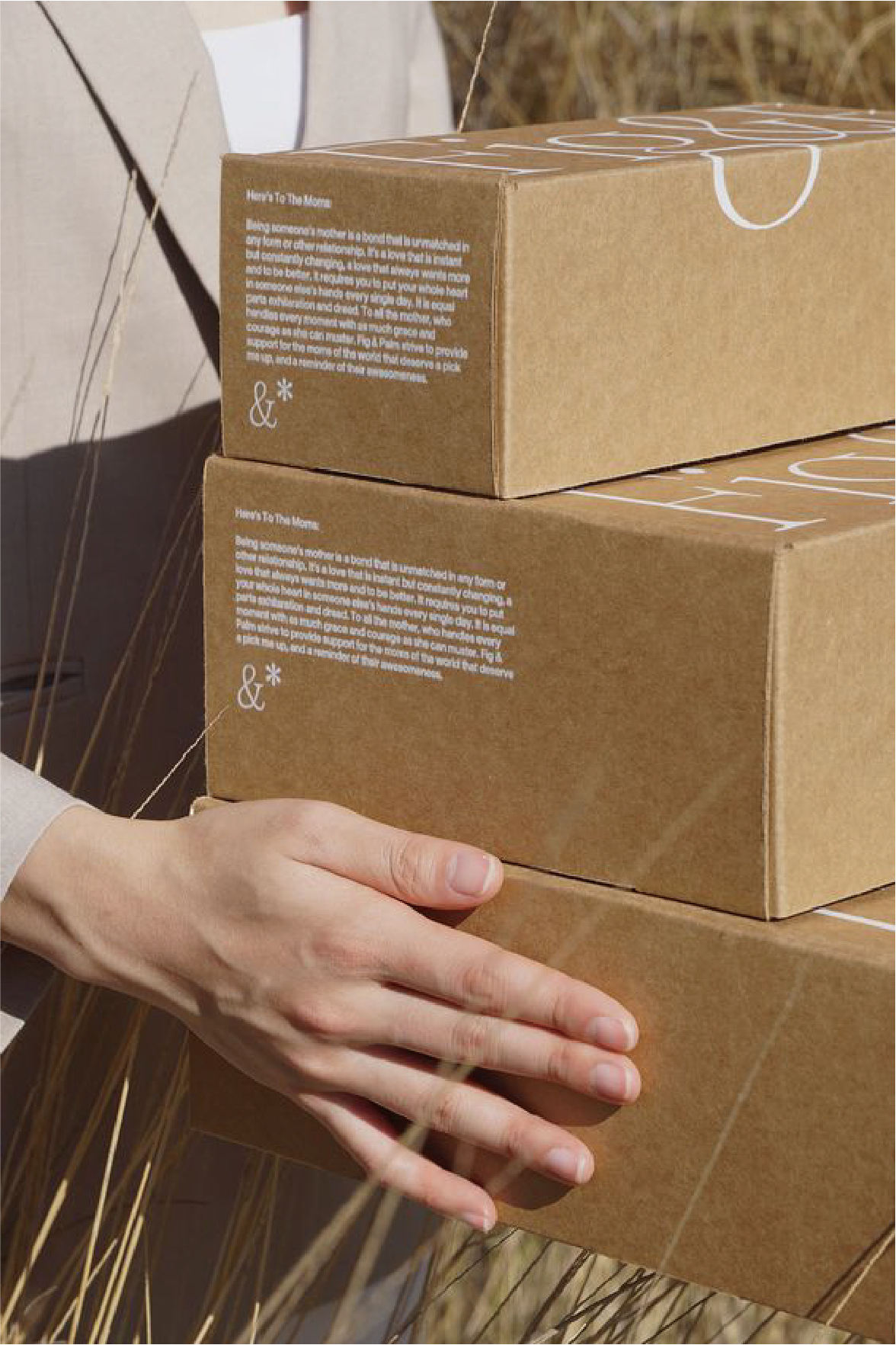 A man holds in his hands two khaki-coloured cardboard boxes with "Here's To The Moms" and a description printed on them.