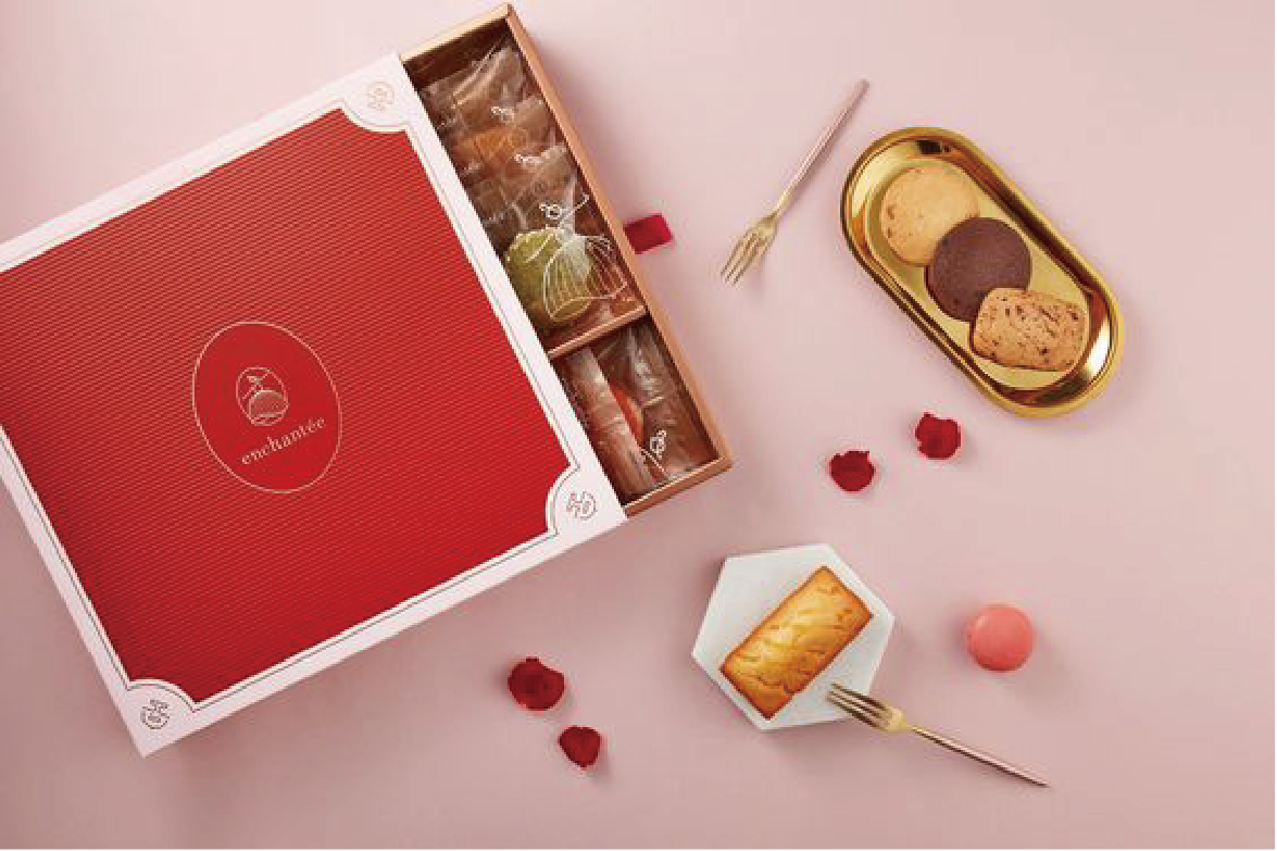 An open gift box containing an assortment of chocolates and pastries, with a gold cutlery piece and a pink round candy next to the box, all set on a pink background.