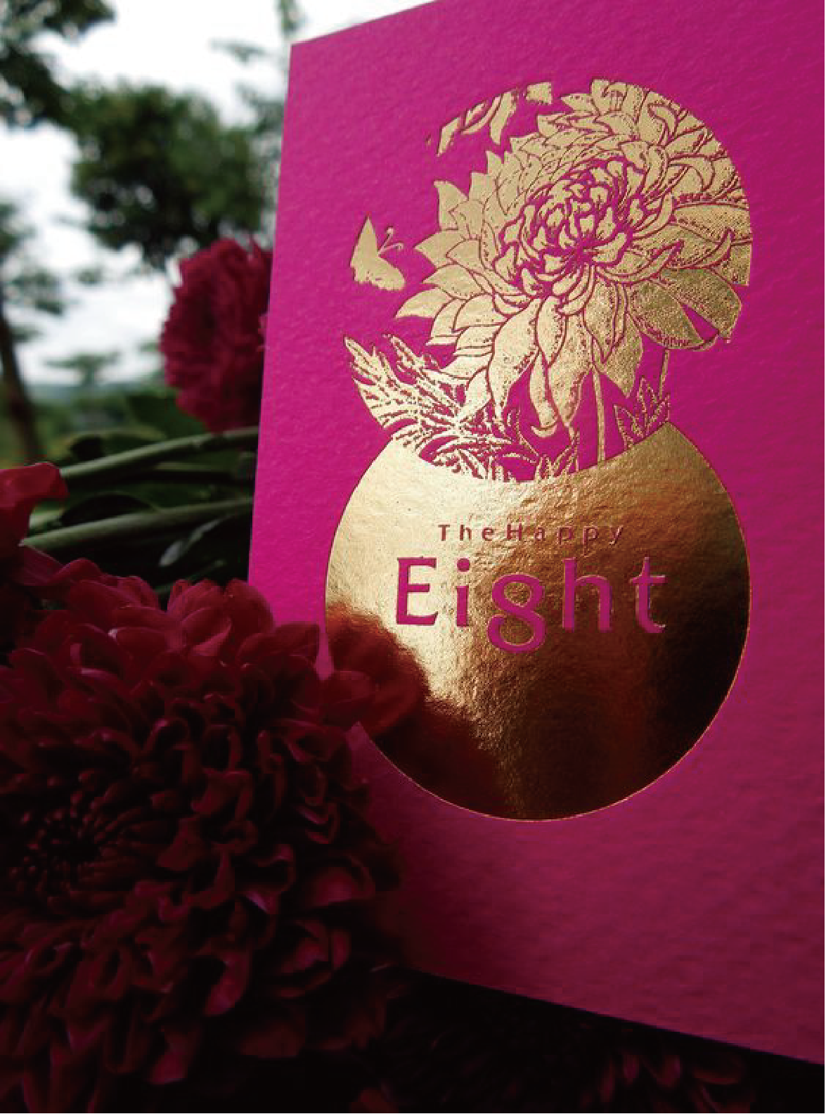 The photo shows a pink greeting card with a gold floral design and butterflies, the floral design is on a gold oval. In the middle of the gold oval are the words "The Happy Eight".