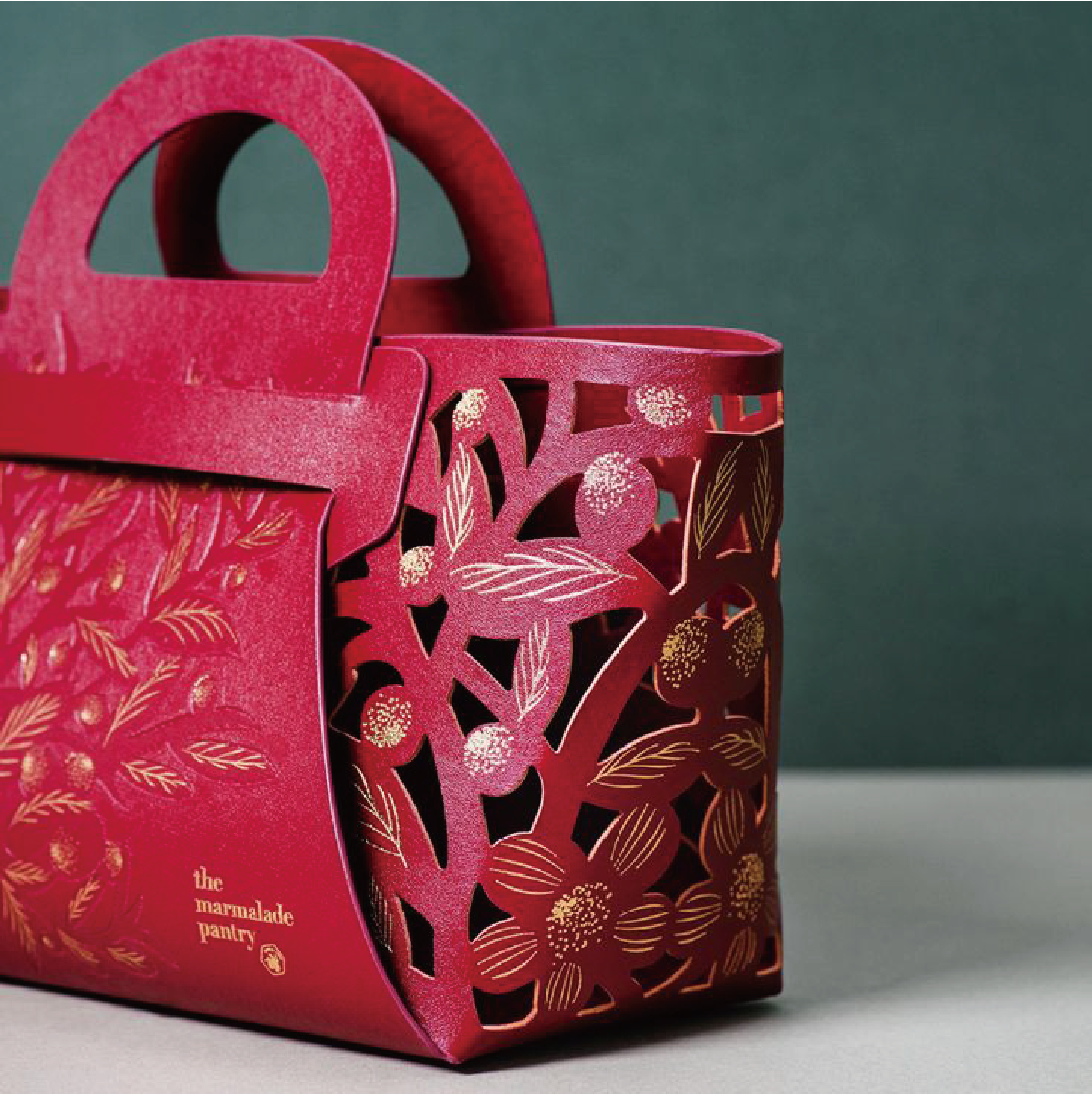 This image shows an ornate dark red carrier bag with a gold botanical and floral openwork design. The words "The Marmalade Pantry" are printed on the side of the bag.