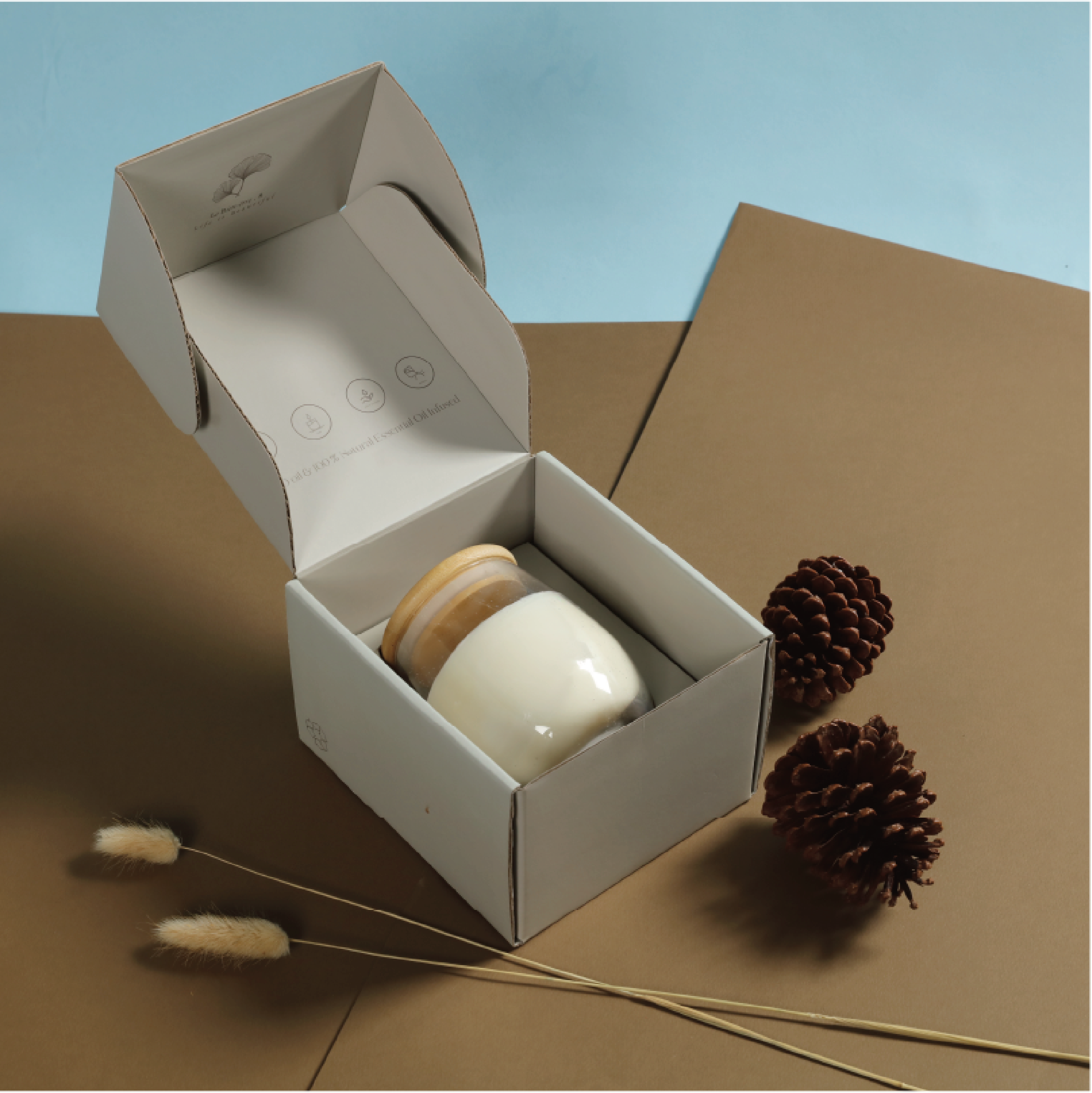 A matt white kraft mailer box, the box is half open and contains a round soap and two wooden soap holders. The background is a light blue and brown cardboard, with pine cones and a few dried plants scattered around.