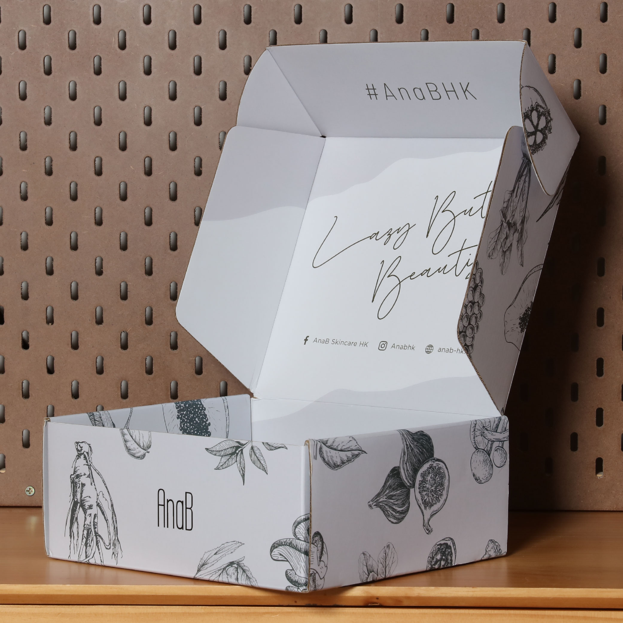 An open matte white card mailer box featuring a hand-drawn black and white illustration of fruits and leaves with the brand's logo 'AnaB'. The brand's social media information is printed on the inside of the box.