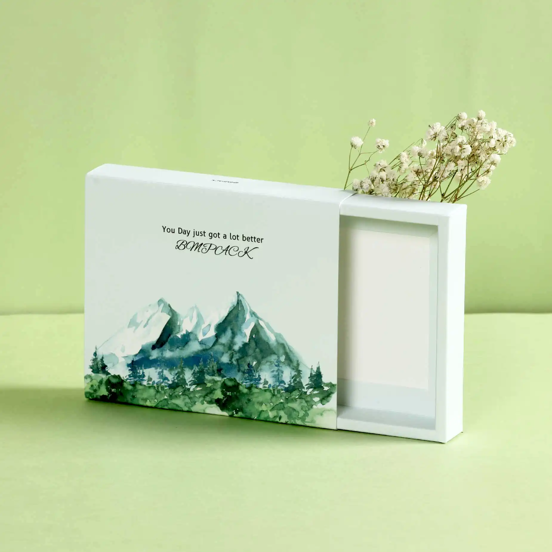 A white box with a watercolour painting of a mountain range and the words "Your Day just got a lot better" printed on the top. Next to the box is a small bouquet of dried white flowers on a light green background.