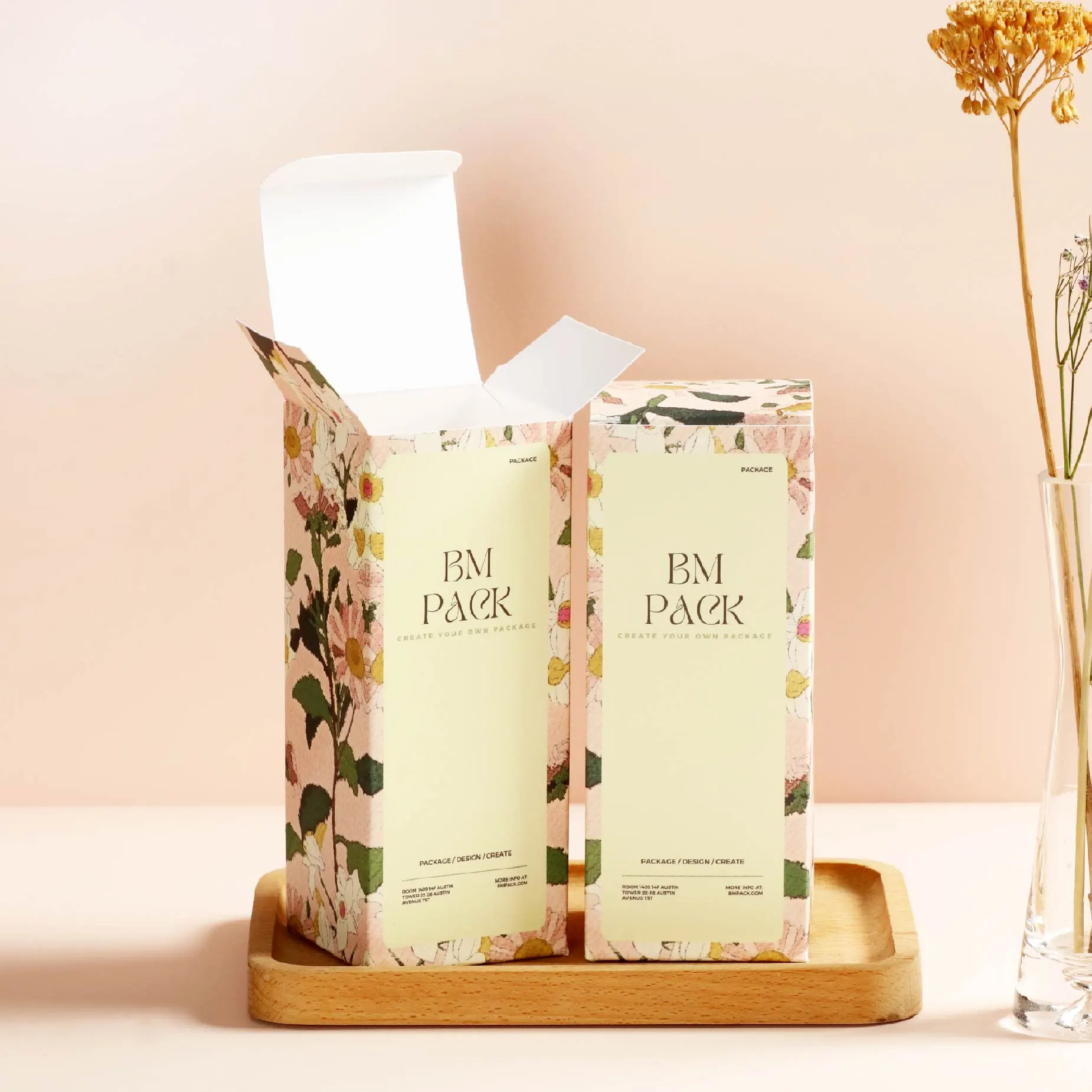 Two BM PACK boxes with a pink and yellow background and floral design with brand and product information printed in the centre. The boxes are placed on a wooden tray with a pink background and dried flowers in a vase next to them.
