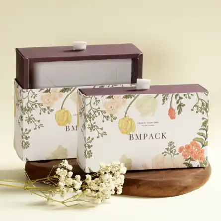 Two BM PACK boxes with floral motifs, one larger and one smaller, with the brand logo printed on the front. The boxes have a light brown handle on top and rest on a round wooden board next to a bunch of dried flowers.