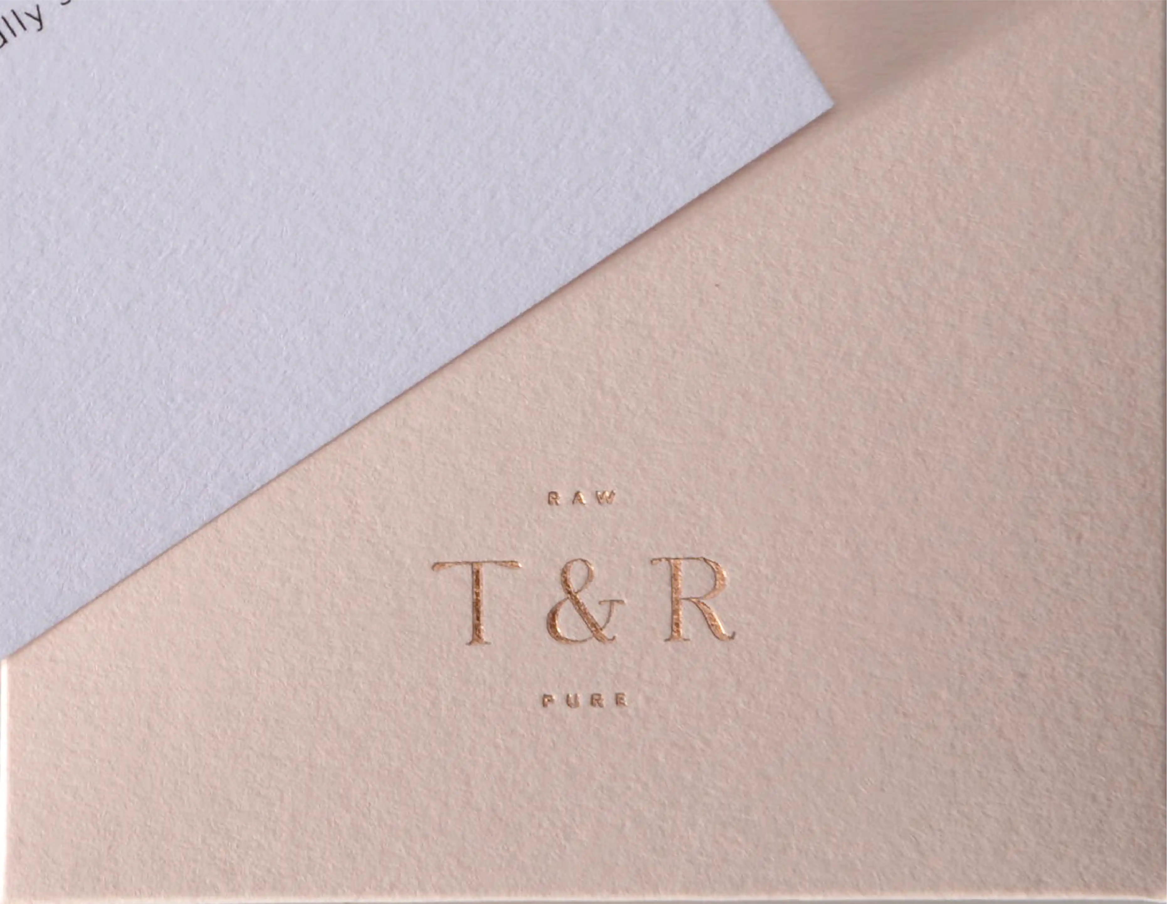 This is a fine piece of textured white cardboard corner, the top half of the paper is white, the bottom half is light brown. The surface of the paper has a unique texture. On the brown area, the words "RAW T&R PURE" are printed in gold.