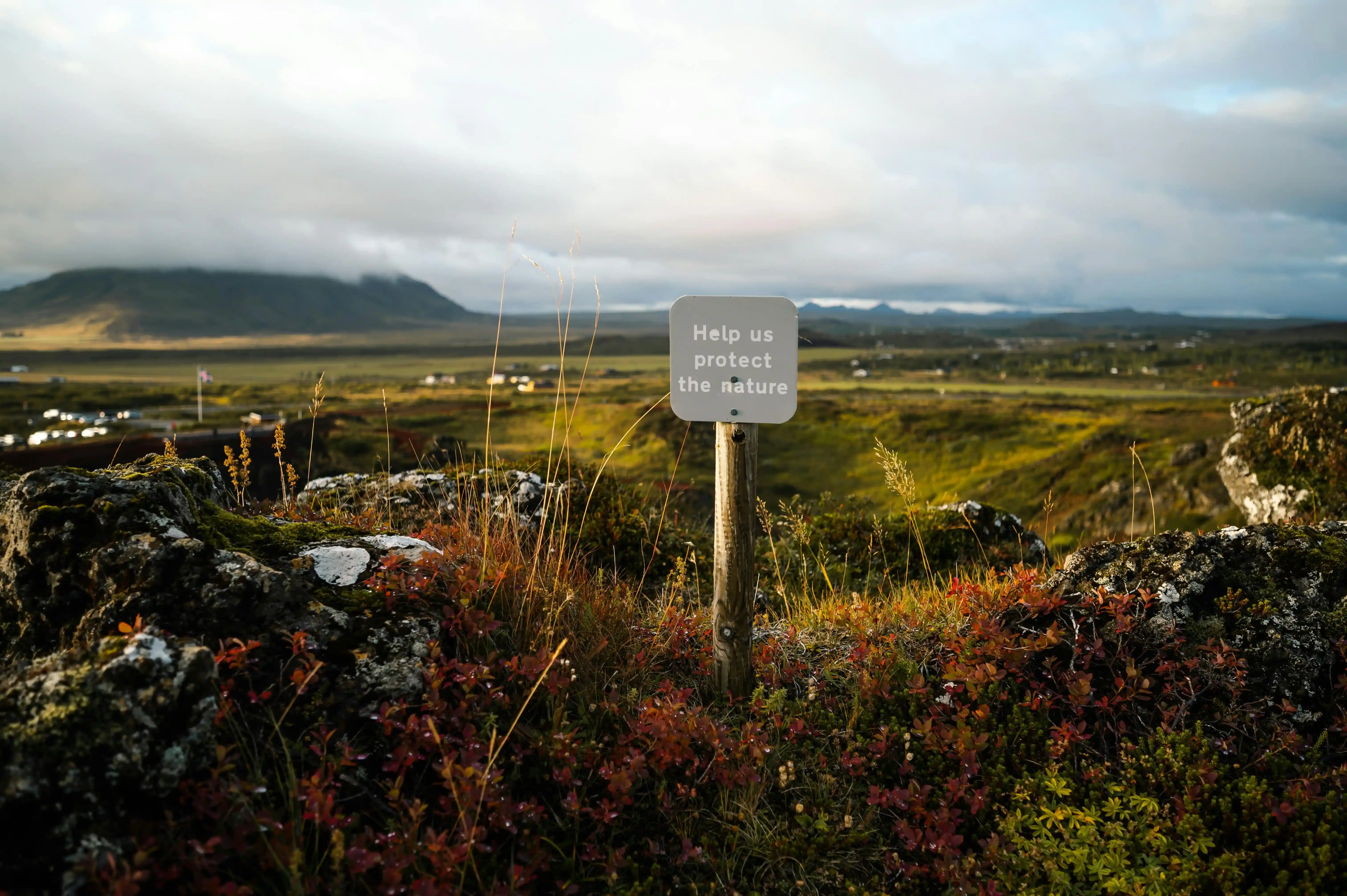 The warning sign "Help us protect the nature" in the natural landscape appeals to the environment and is mirrored by the mountains and sky in the distance.