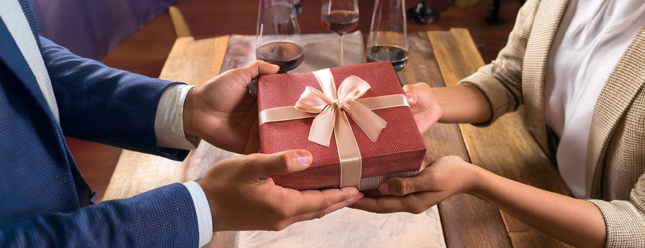 Gift Bags: Essential Tips to Enhance the Gifting Experience; a man in a suit presents a red gift tied with a gold bow to a woman sitting across from him